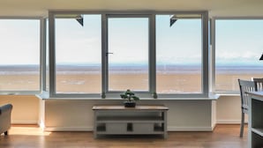 A room with a large window overlooking a beach. There is a small wooden bench with a plant on it, and a chair is visible to the right.