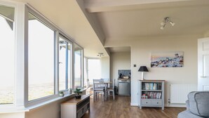 A spacious living area with large windows, a dining table, a kitchen area, and a bookshelf.
