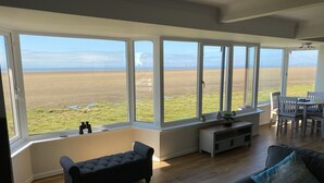 A spacious living room with large windows, a dark grey sofa, a wooden floor, and a dining area with a table and chairs.