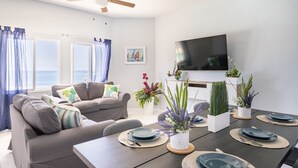 A modern living room with a sofa, dining table set for four, and a flat-screen TV mounted on the wall.