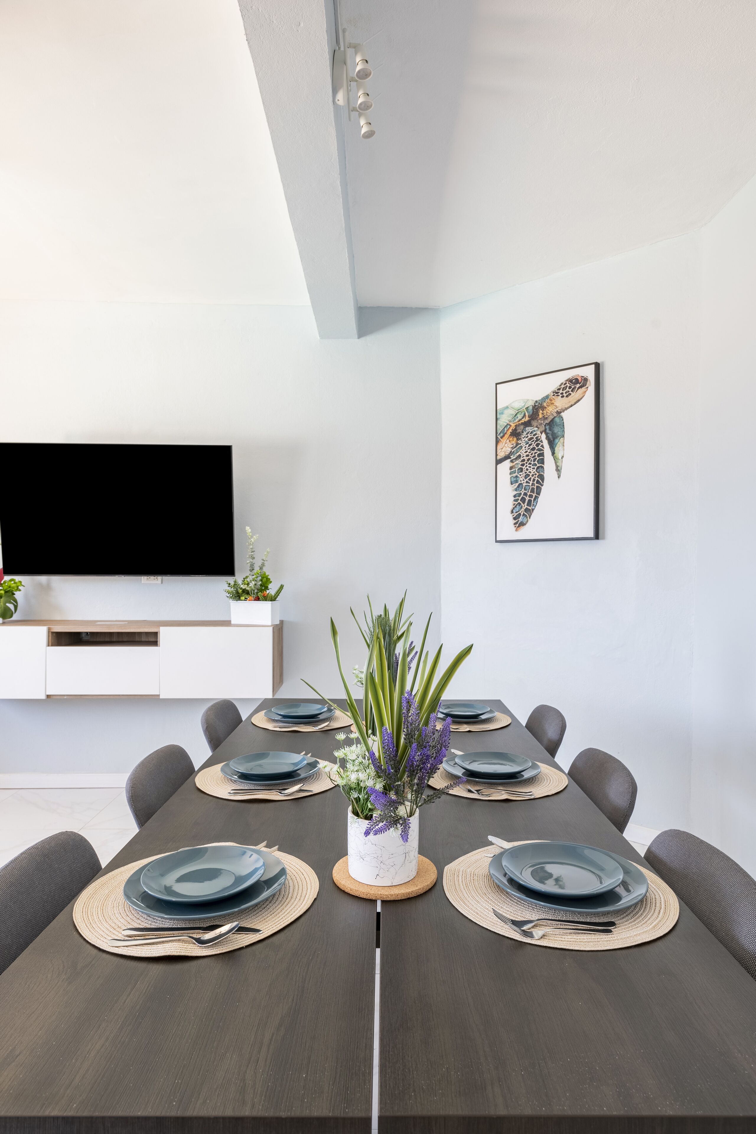 A modern dining area with a dark wood table set for six, white cabinetry, a flat-screen TV, and a framed sea turtle artwork on the wall.