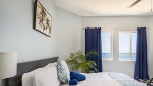 A neatly made bed with a white and patterned bedspread, a grey headboard, and two blue towels. There is a large window with blue curtains, a potted plant, and a painting on the wall.