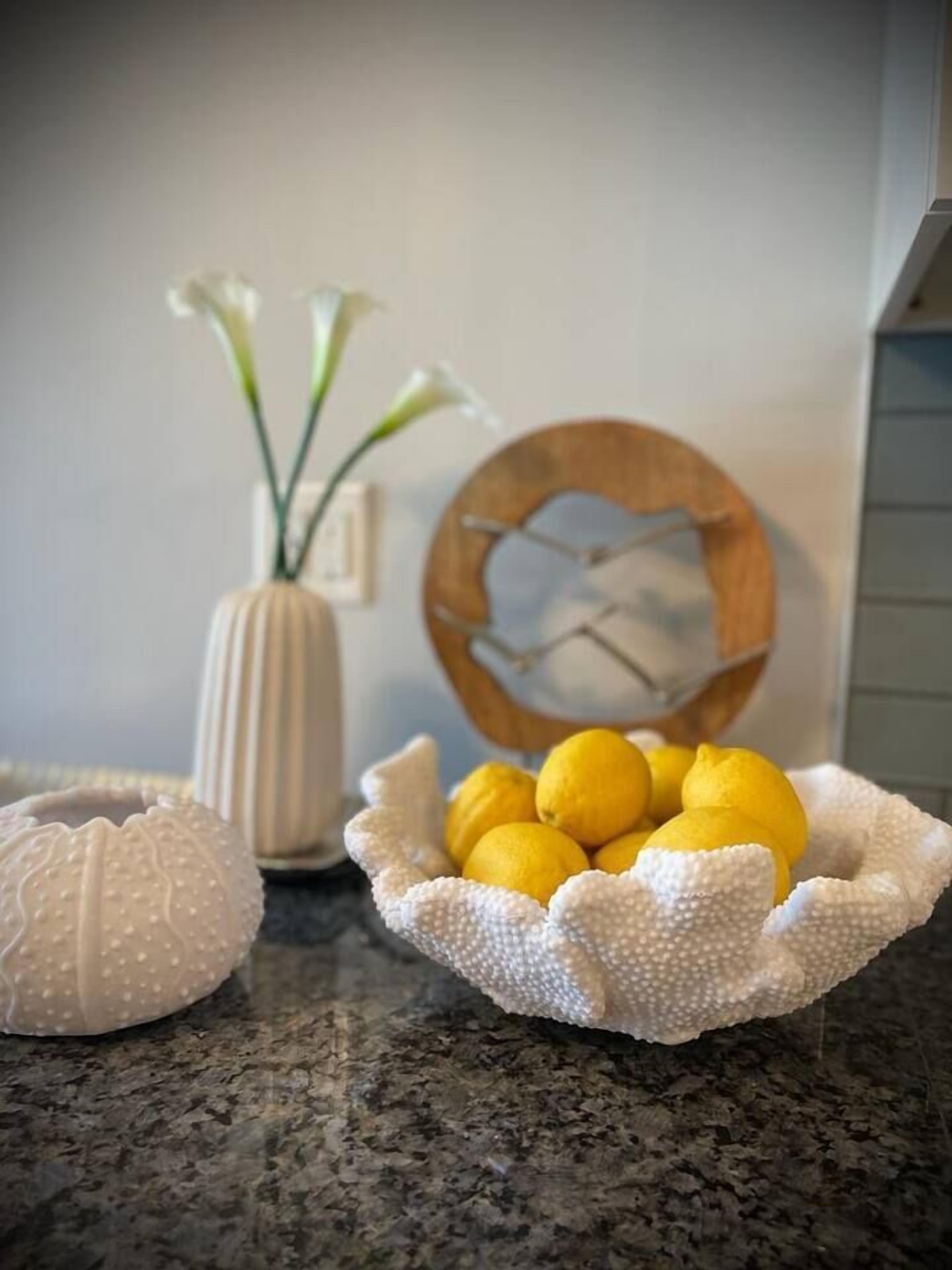 A kitchen counter with a bowl of lemons, a white vase with flowers, and a wooden clock.