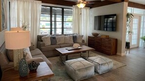 A spacious living room with a wooden ceiling, a large window with curtains, a sofa, a coffee table, and a television mounted on the wall. - 04 Rustic meets beachy in Wilton Manors! (Wilton Manors)