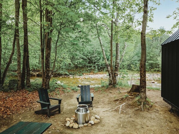 A forested campsite with two chairs, a portable stove, and a water container.