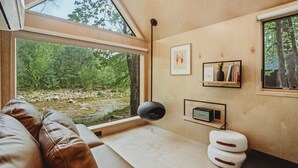 A modern room with a large window, a black floor lamp, a flat-screen TV, and a small shelf with books and a plant. - Lumen Nature Retreat | Cabin HYGGE-ASTRID (North Woodstock)