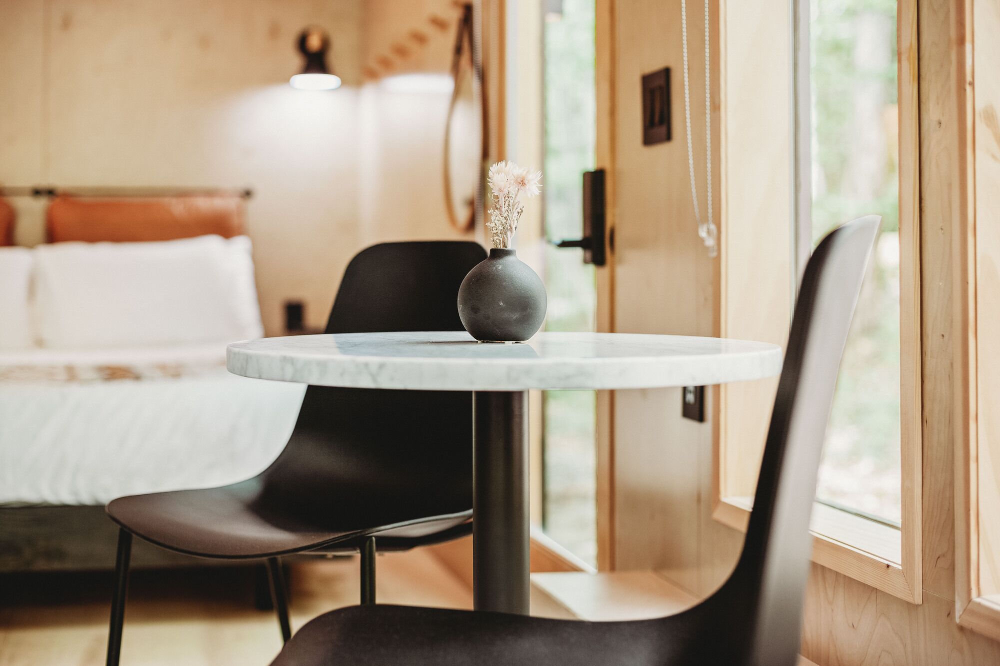 Quarto moderno com mesa redonda, duas cadeiras pretas, um vaso com flores e uma cama com roupa de cama branca.