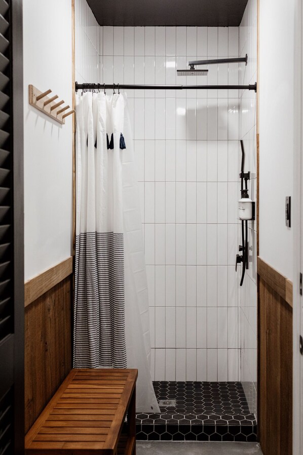 Un petit espace de douche fermé, avec un banc en bois, des murs recouverts de carreaux blancs et un rideau de douche à rayures noires et blanches.