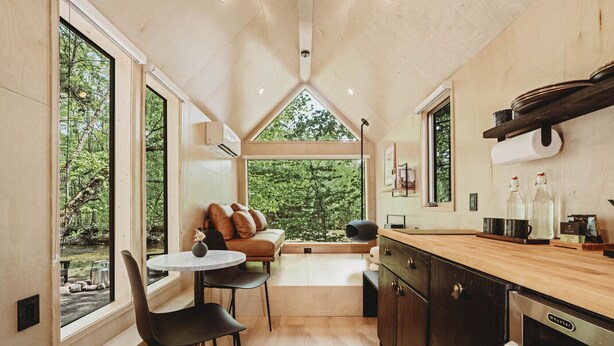 Cocina compacta con encimera de madera, un lavamanos y lavavajillas. Hay una zona de comedor con mesa y sillas, y un sofá. Amplios ventanales ofrecen vistas al exterior.