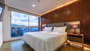 A modern bedroom with a large bed, wooden paneling, a nightstand with a lamp, and a view of the city through large windows. - Stunning views from 25th-floor condo! El Poblado! (Medellín)