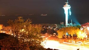 Vista nocturna de una torre con una luz en la cima, rodeada de árboles y coches estacionados.