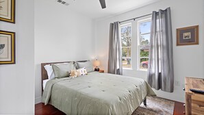 A bedroom with a large bed, a bedside table with a lamp, a dresser, and a window with curtains.