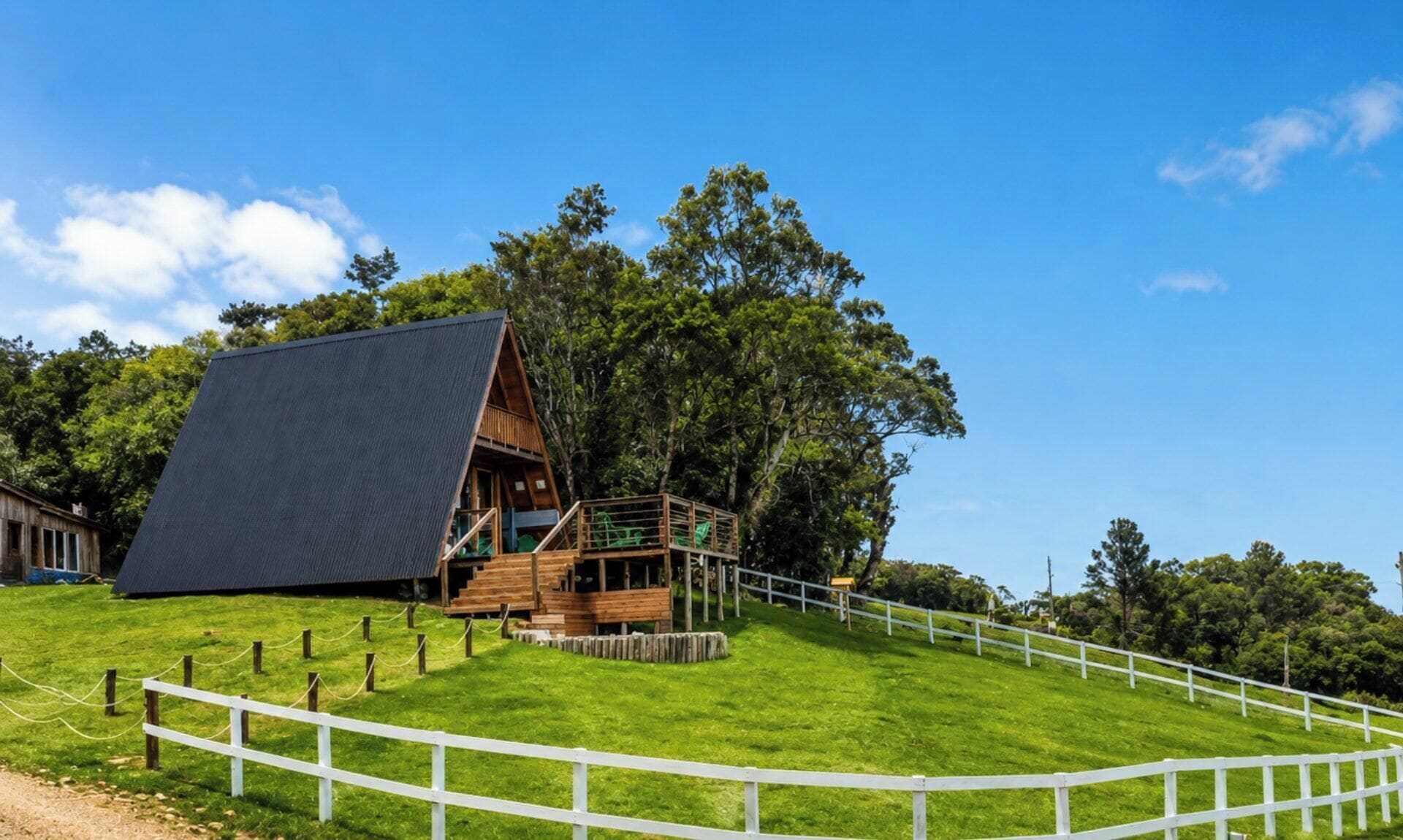 Una cabaña de madera con techo inclinado, rodeada por una cerca blanca, ubicada en un área verde con árboles al fondo.