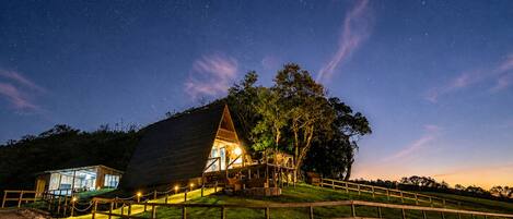 A wooden cabin with a glass facade, surrounded by a wooden fence, set against a starry night sky.