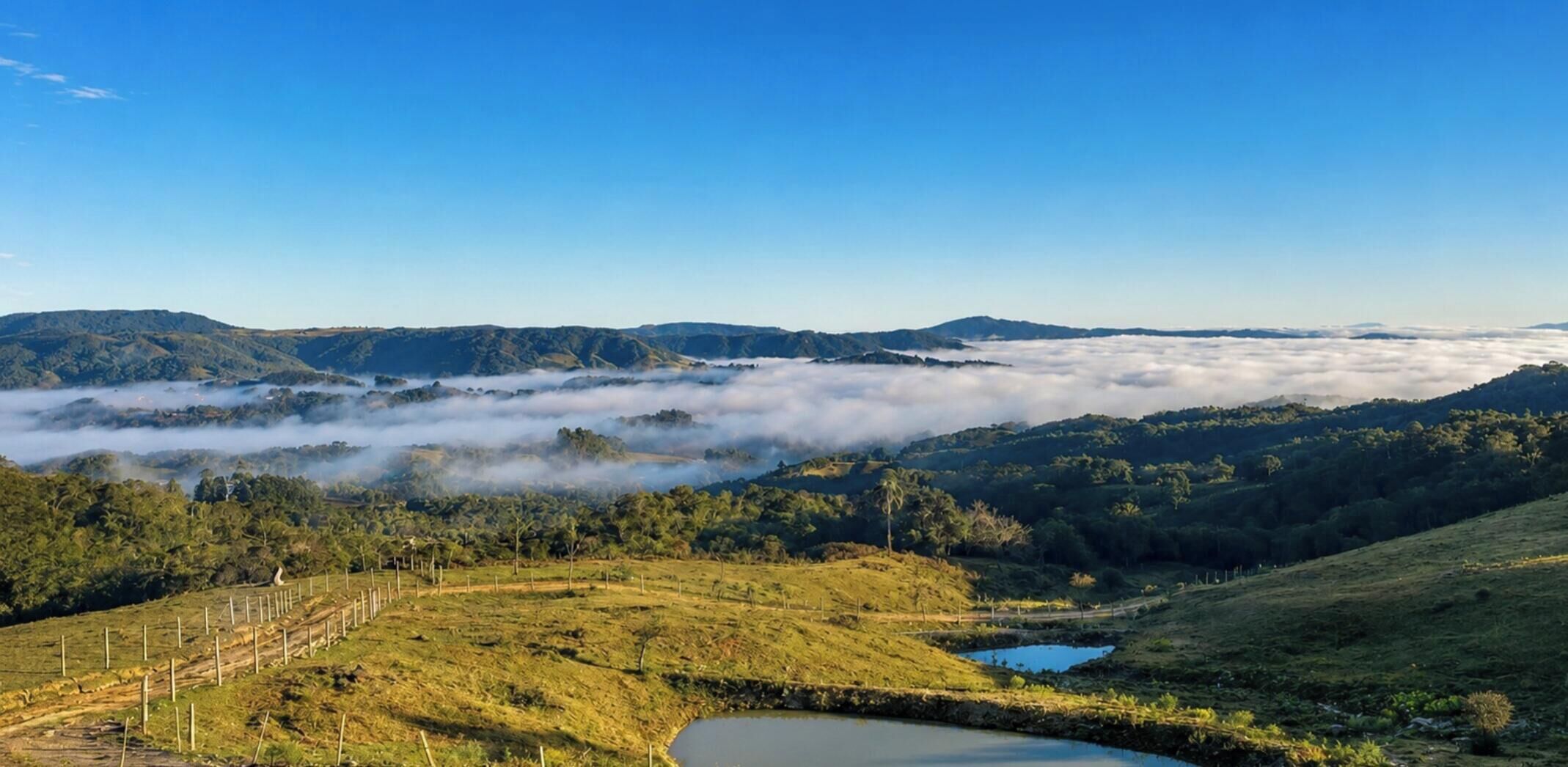 A panoramic view of a hilly landscape with a clear sky, a body of water, and a fence line.