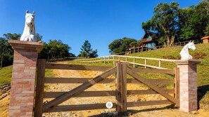 A gated entrance with horse statues, a wooden fence, and a hill with trees and a building in the background. - Rancho Sol Poente - Rancho Queimado (Rancho Queimado)