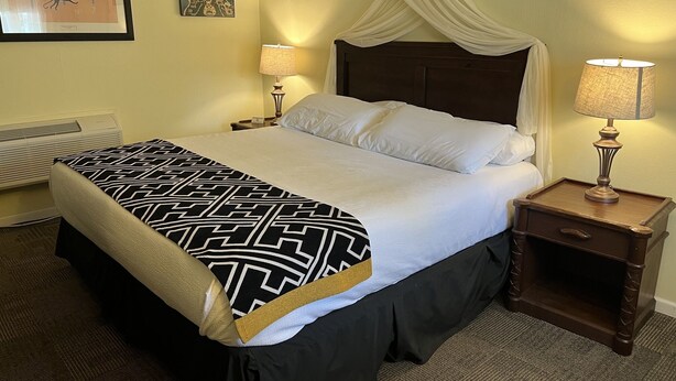 Un lit bien fait, recouvert d’une couvre-lit à motifs noir et blanc, avec deux lampes de chevet et une table de chevet en bois foncé dotée d’un tiroir.