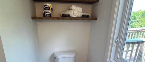 Et lille badeværelse med toilet, skraldespand, et vindue og to træhylder over toilettet.