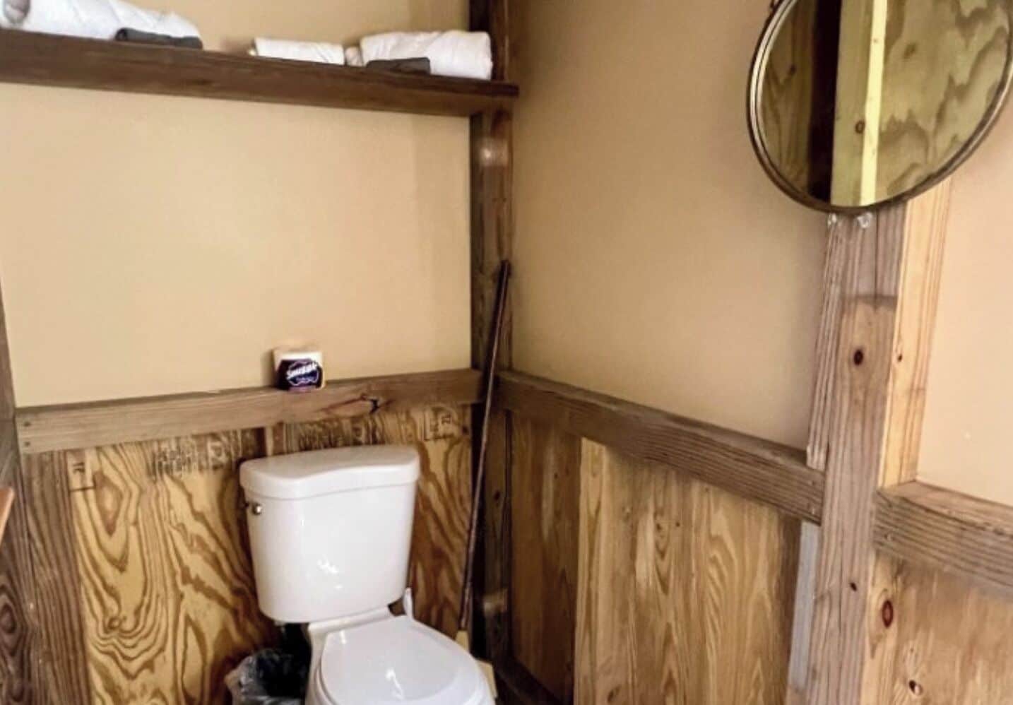 A small, wooden-paneled bathroom with a toilet, a shelf above the toilet holding towels, and a mirror on the wall.
