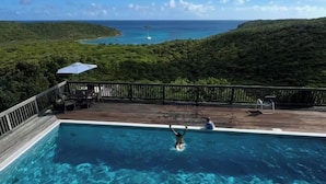 Una piscina con agua cristalina azul, rodeada por una terraza de madera y vistas al océano y las colinas.