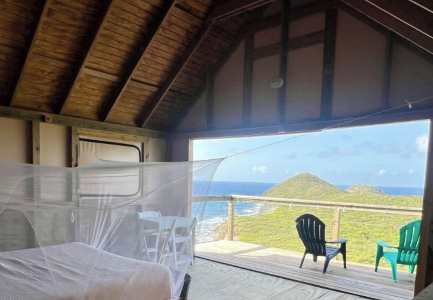 A room with a bed, a table, and chairs, and a balcony with a view of the ocean and hills.