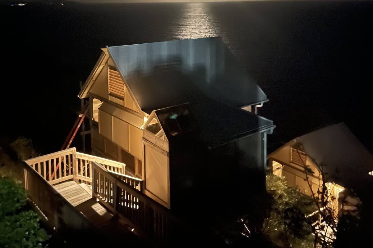 A house with a balcony overlooking a body of water, under a cloudy sky with the moon visible.