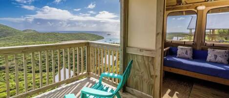 Une terrasse en bois offrant une vue sur un paysage côtier, un canapé bleu et deux fauteuils turquoise.