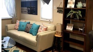 Ein Wohnzimmer mit einem beigefarbenen Sofa, gemusterten Kissen, einem Glastisch, einem Holzregal und einem Teppich mit Muster.
