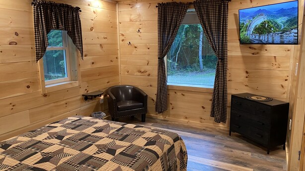Ein Holzhüttenraum mit einem Bett, einem Sessel, einem kleinen Tisch, einem Fernseher und einem Fenster mit Vorhängen.
