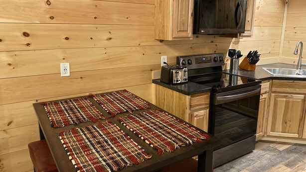 A kitchen with wooden cabinets, a black microwave, a stove, and a table with a woven placemat. - Bobcat Corner, Hot Tub, Secluded, Pet Friendly (Logan)