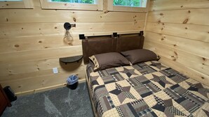 A wooden cabin bedroom with a bed, two windows, a nightstand, and a wall-mounted lamp. - Bobcat Corner, Hot Tub, Secluded, Pet Friendly (Logan)
