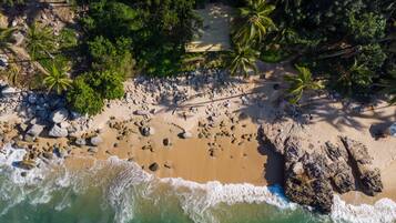 An aerial view of a beach with a wooden walkway, palm trees, and rocky shoreline.