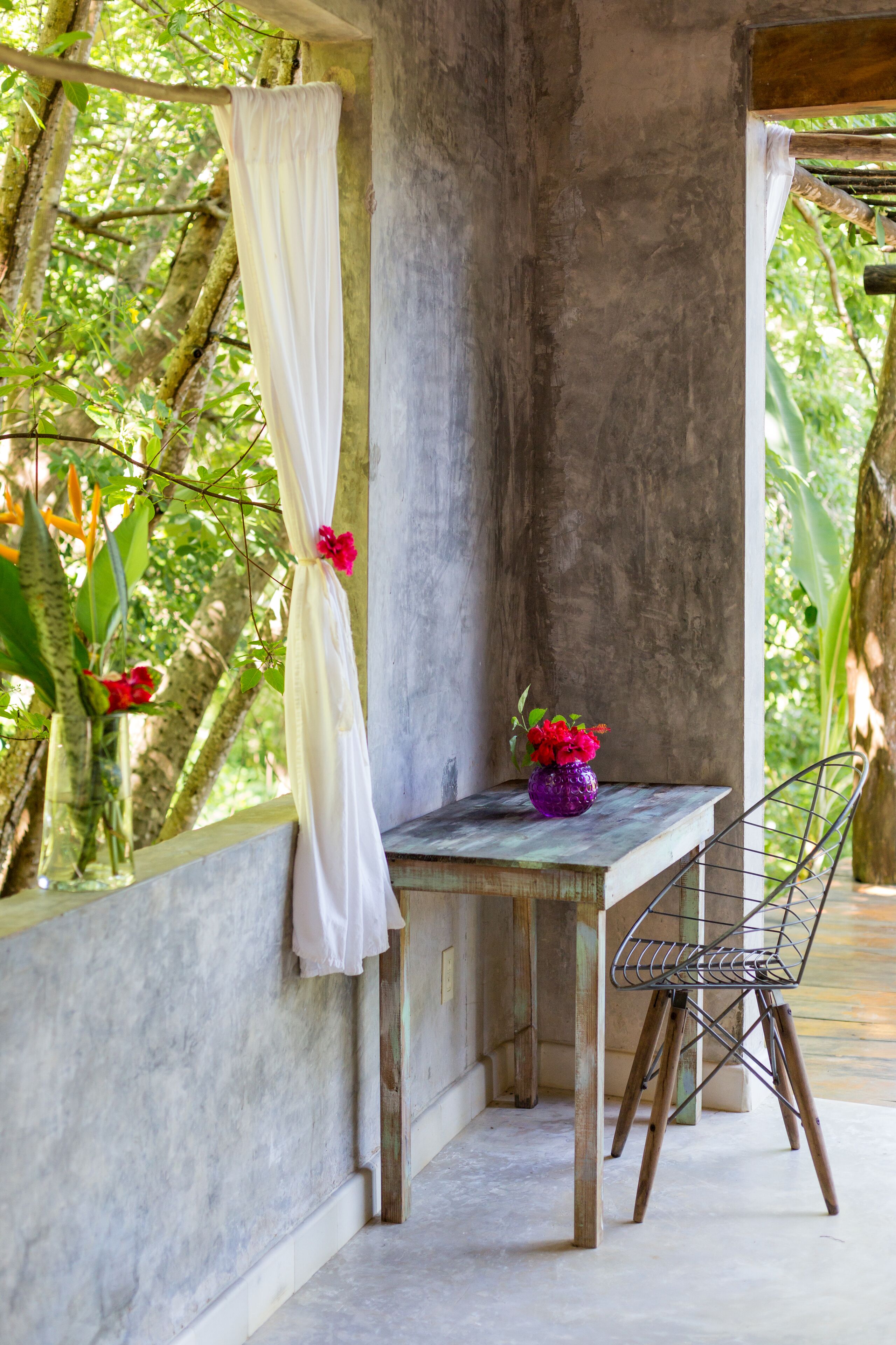 A rustic wooden table with a metal chair, a white curtain, and a vase with red flowers against a concrete wall, surrounded by greenery.