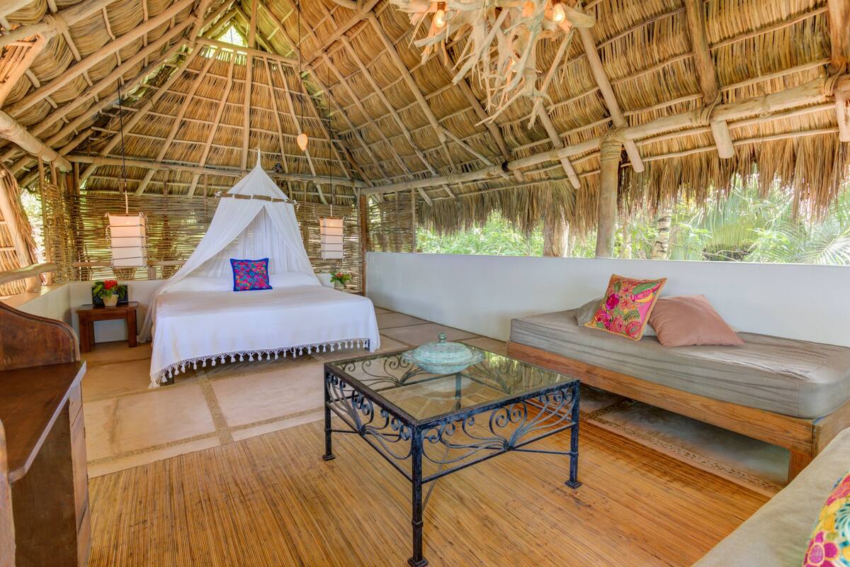 Una habitación con techo de paja, una cama, un sofá y una mesa de centro de vidrio.