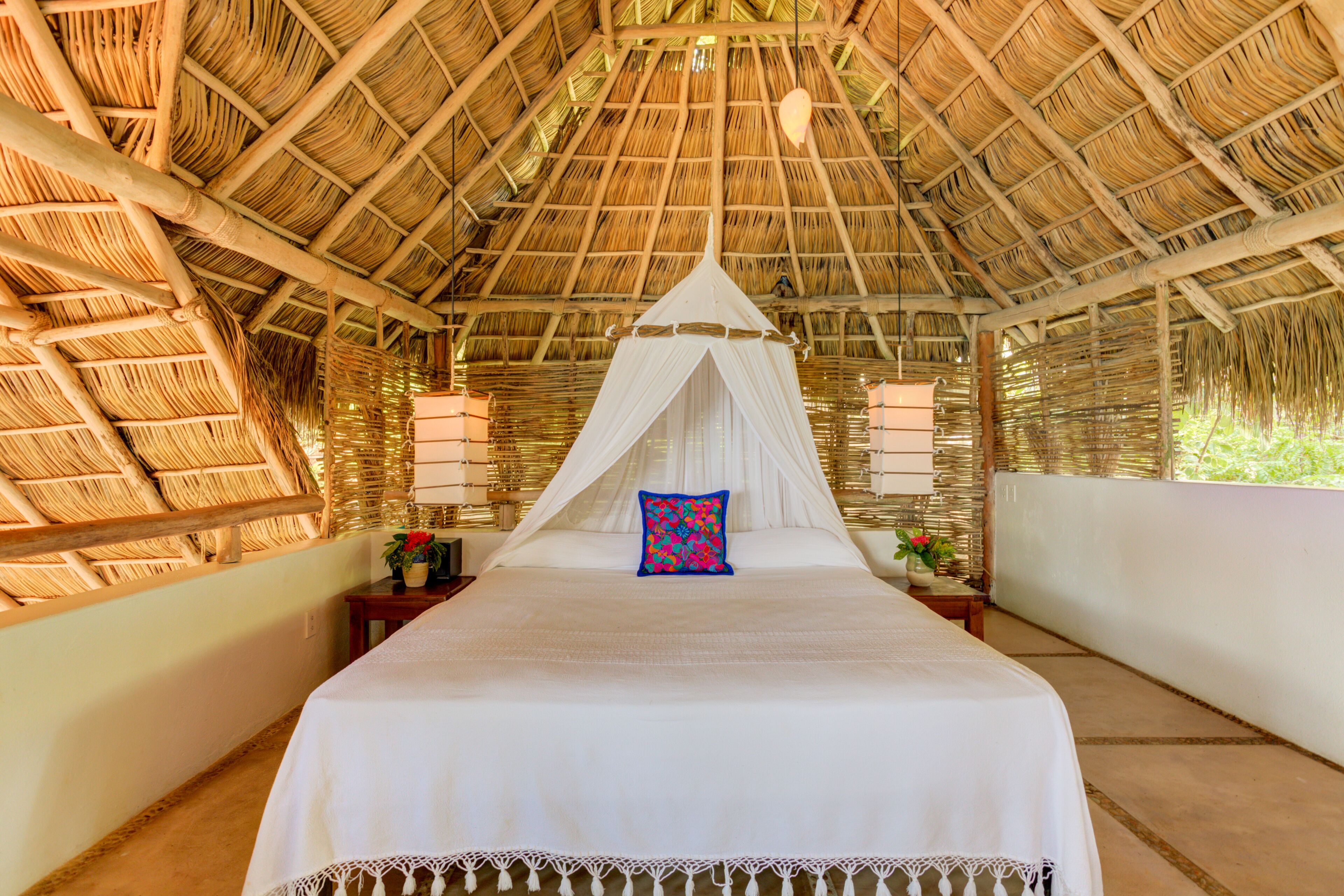 A room with a bed under a thatched roof, a white canopy, and a nightstand with a lamp.