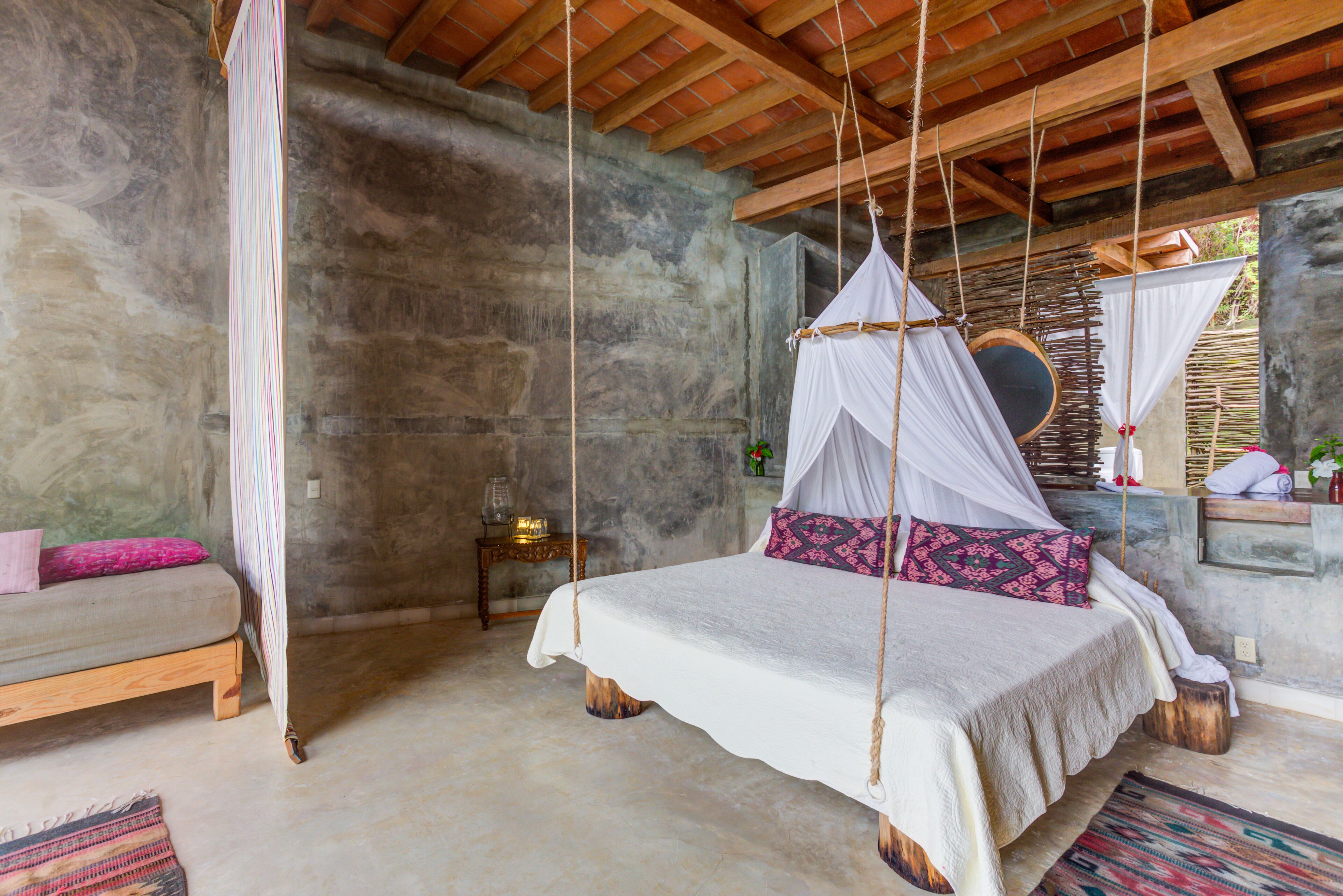 A bedroom with a bed, a bench, a hanging chair, and a stone wall.