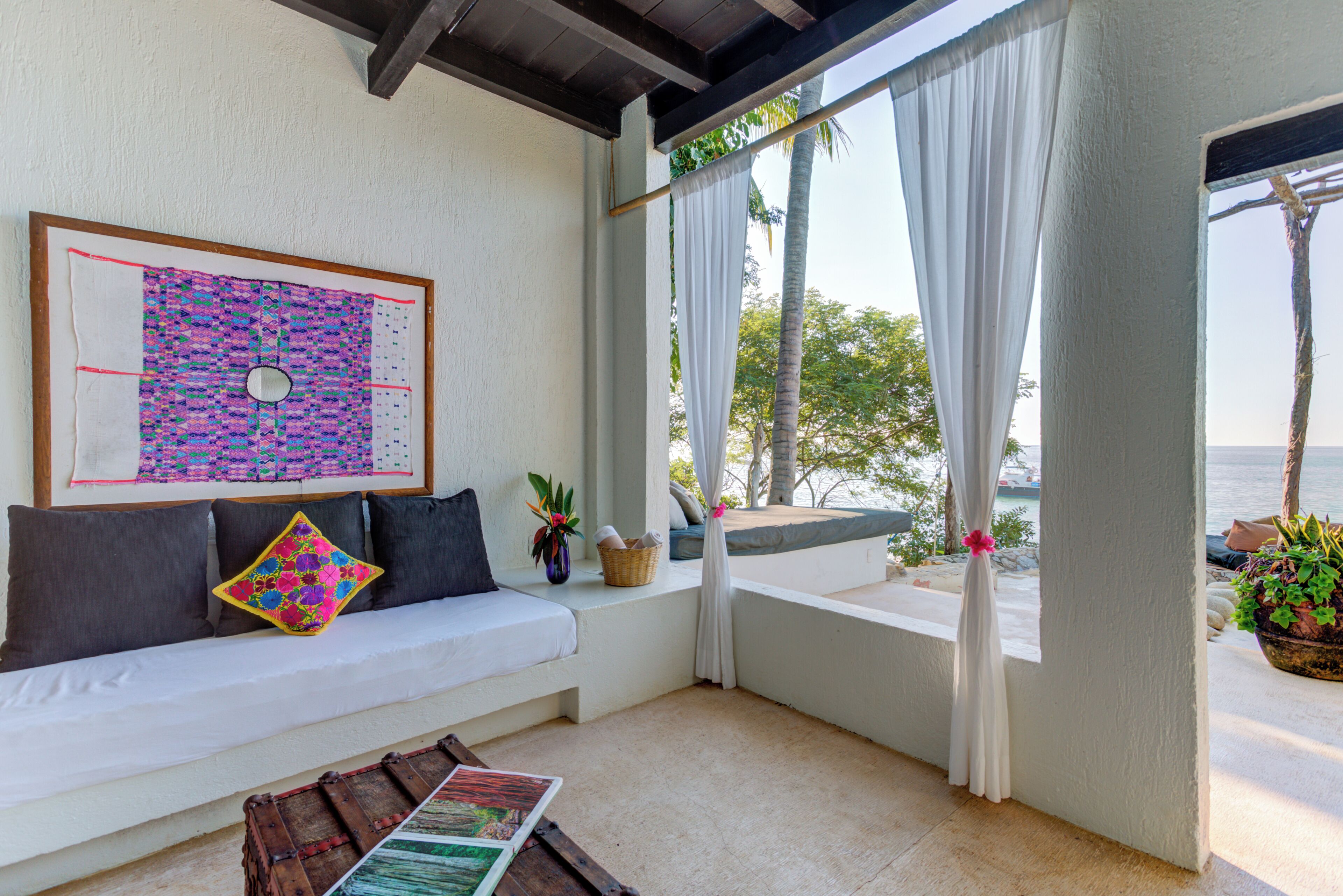 A room with a white sofa, a framed artwork, a wooden table, and a view of the sea through sheer curtains.