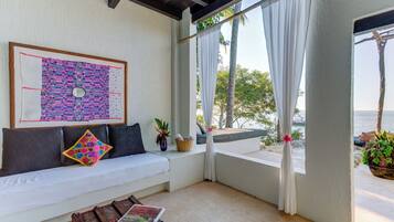 A room with a white sofa, a framed artwork, a wooden table, and a view of the sea through sheer curtains.