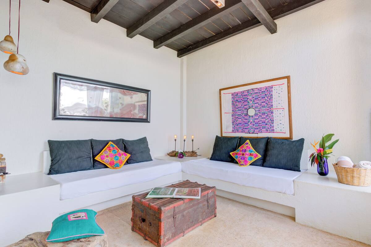 Un espacio de sala acogedor con un sofá blanco, almohadones decorativos, un cofre de madera y arte en la pared.