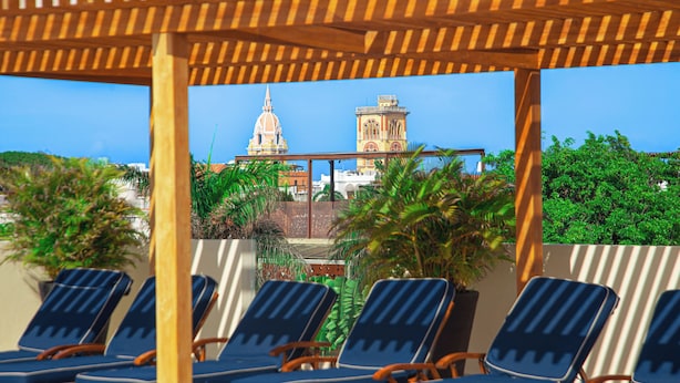 A rooftop lounge area with wooden pergola, blue lounge chairs, and views of a historic building with a dome and tower. - San Diego 974 Suites (Cartagena)