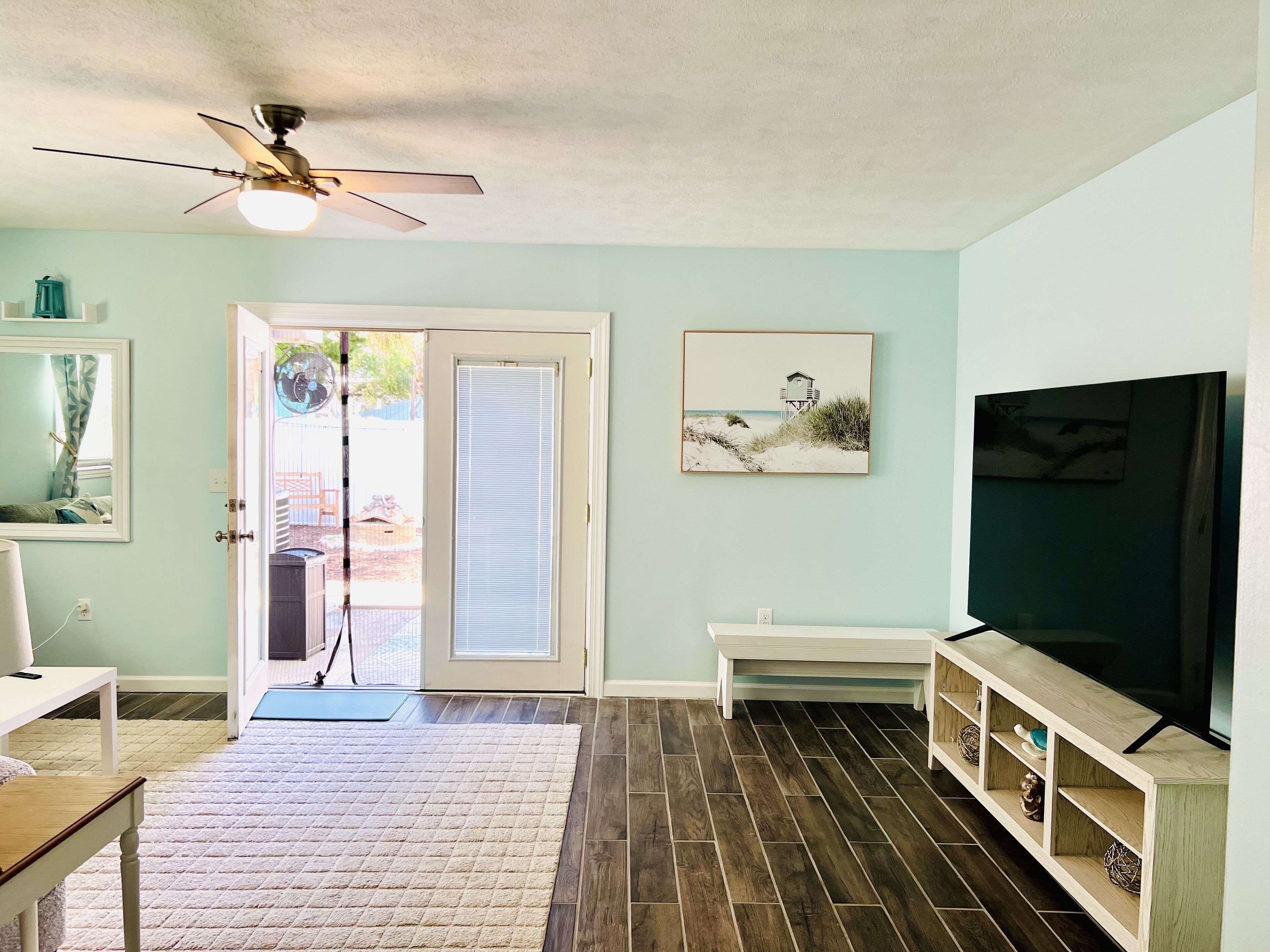 A living room with a ceiling fan, a TV on a wooden stand, a door leading outside, and a rug on the floor.