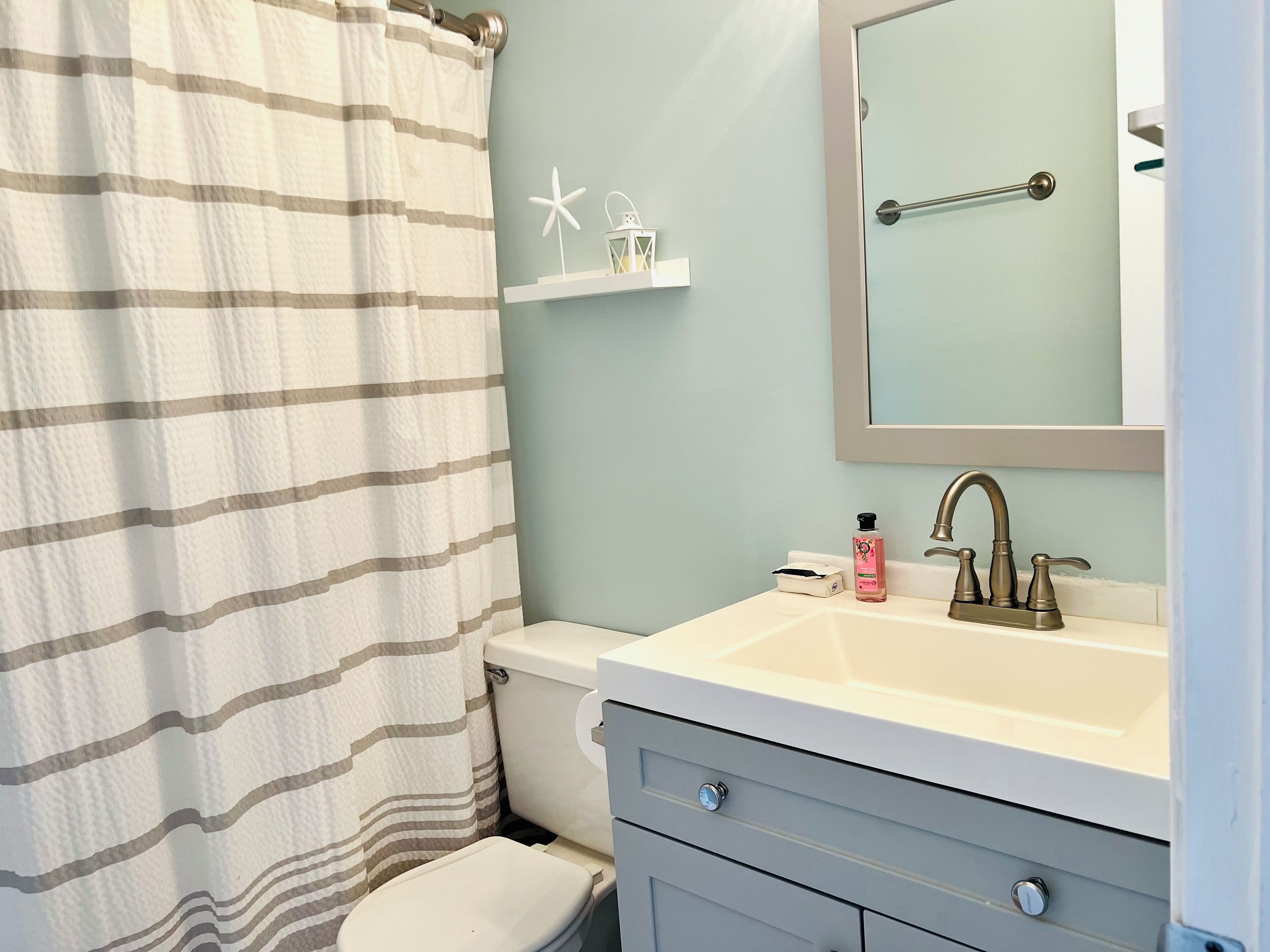 A bathroom with a striped shower curtain, a toilet, a sink with a mirror, and a small shelf with decorative items.