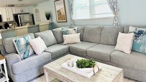 Un soggiorno moderno con un divano modulare grigio, un tavolino in legno con una pianta, e una cucina sullo sfondo.