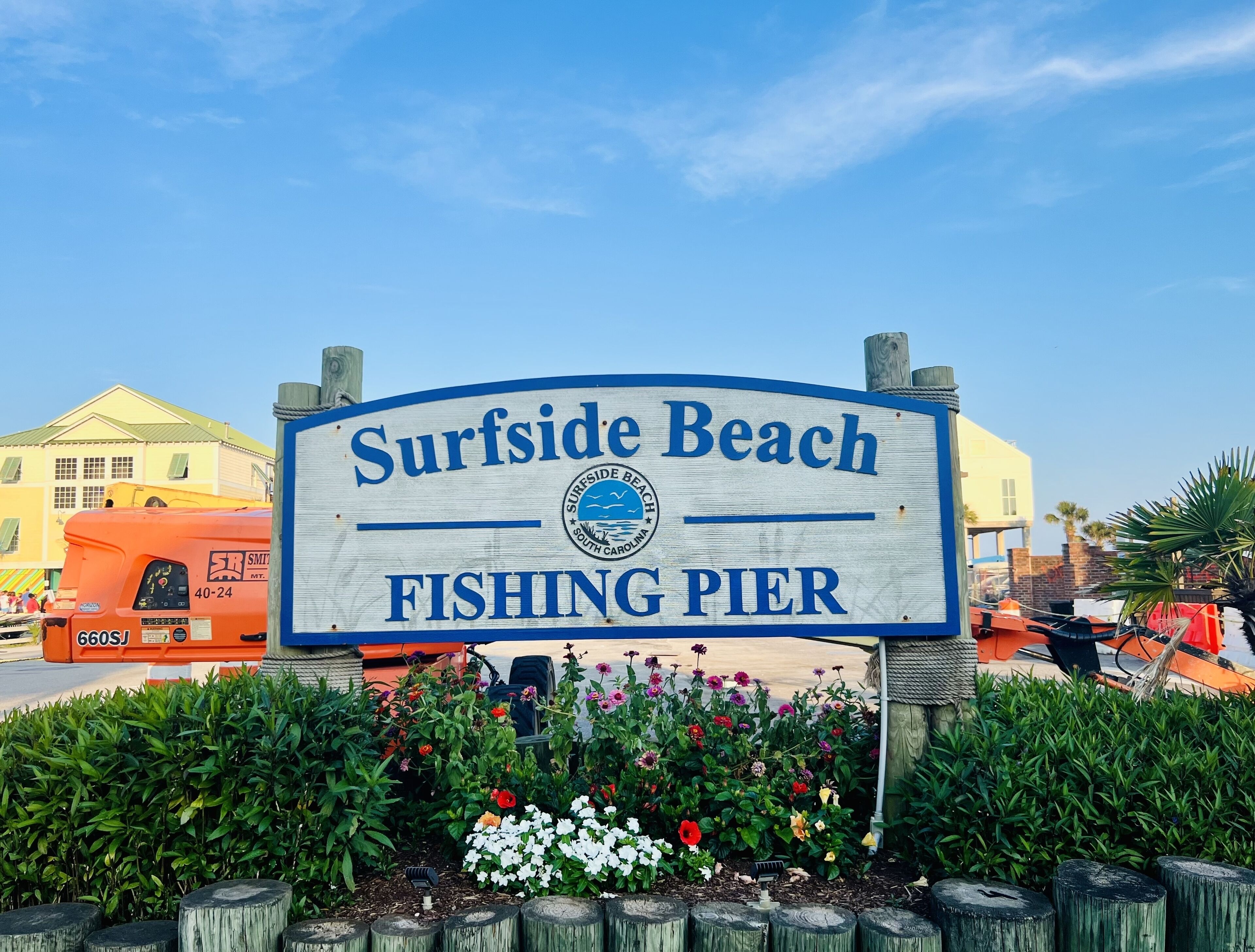 A sign for Surfside Beach Fishing Pier with a logo, surrounded by flowers and wooden posts.