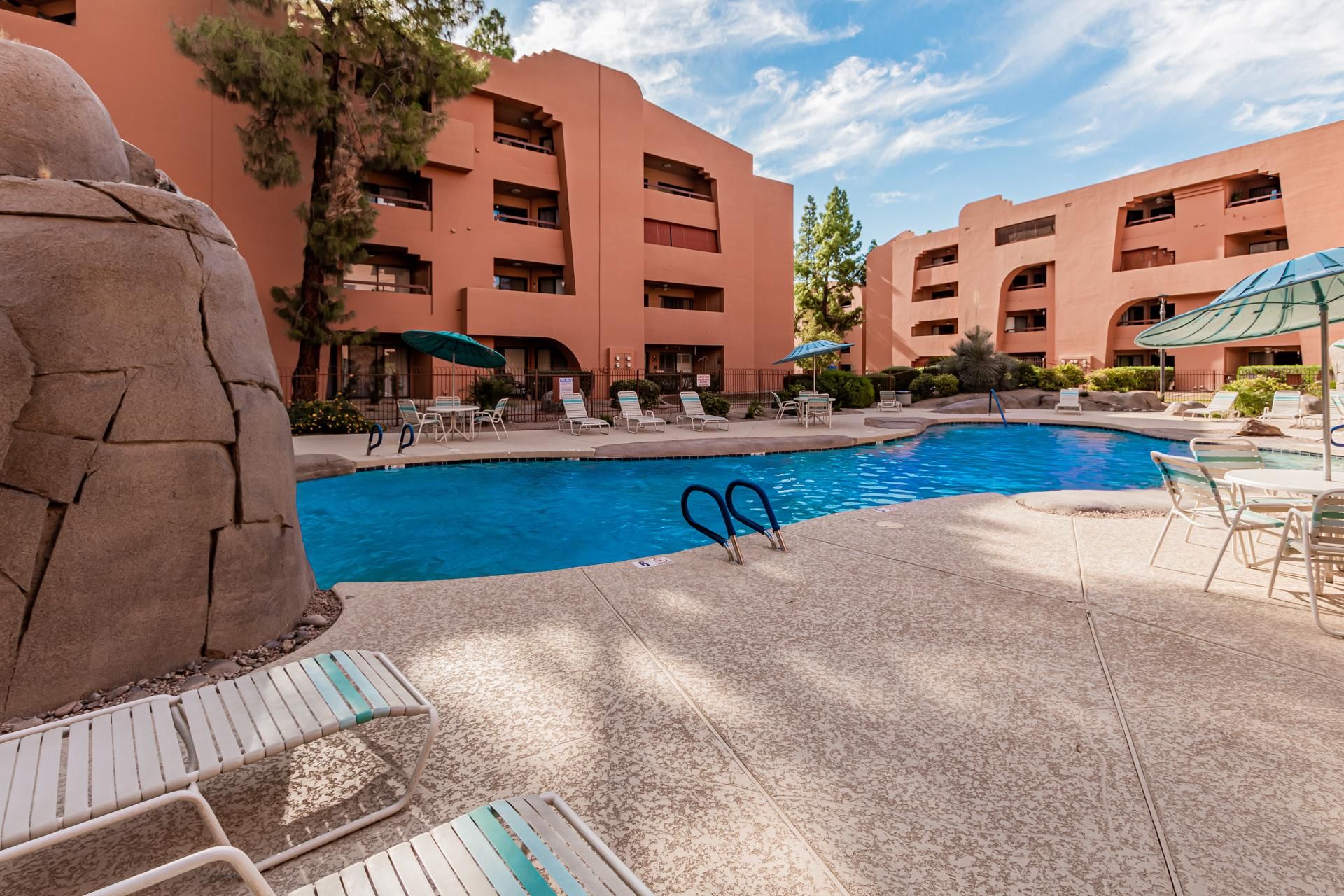 Outdoor pool area with textured concrete deck, white lounge chairs, metal tables, and blue umbrellas.
