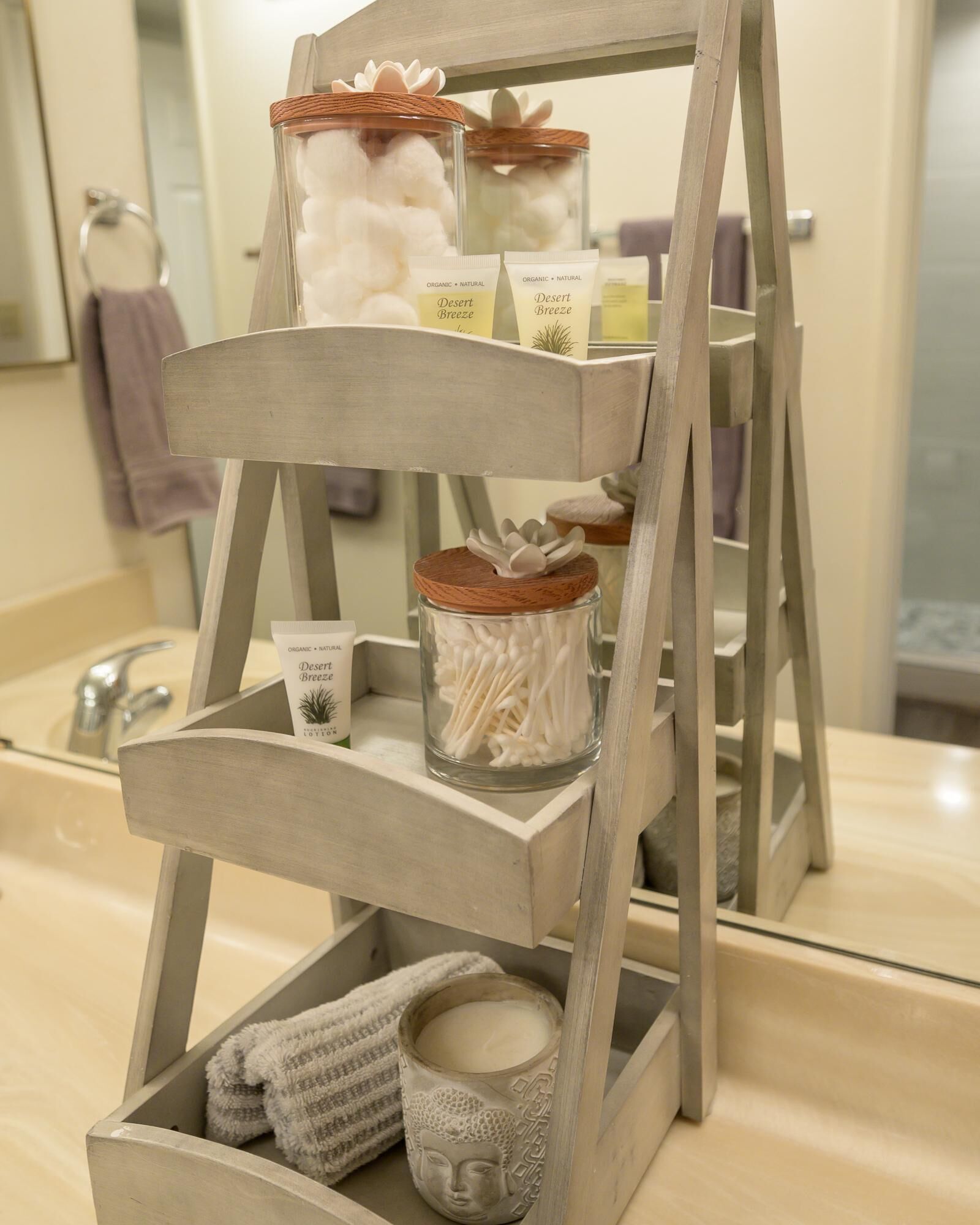 A wooden three-tiered shelf with jars containing cotton swabs and a candle, placed on a bathroom counter.