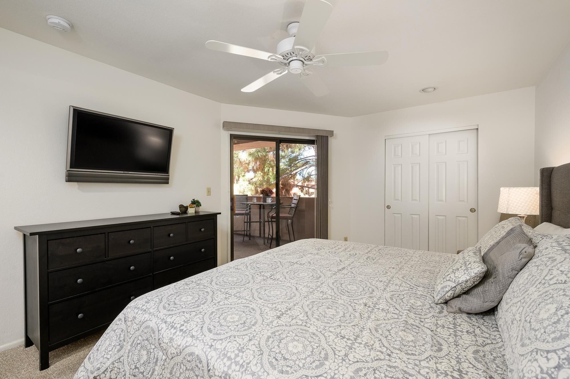 A bedroom with a bed, dresser, TV, ceiling fan, and a door leading to a balcony.
