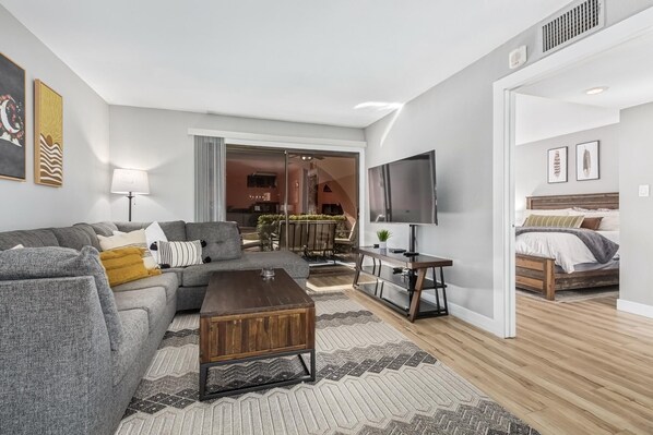 A modern living room with a sofa, coffee table, and TV. There is a bed with a wooden headboard and a small table with a plant. - CASA T'OSO-Anasazi Village-1st floor pet friendly (Phoenix)