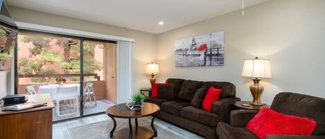 A living room with a sofa, coffee table, and lamps. There is a painting on the wall and a view of a balcony with plants.