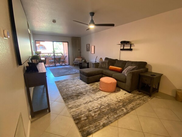 A living room with a sectional sofa, ottoman, and a ceiling fan. There is a dining area visible through a glass door. - CASA AVENTURA - Anasazi Village - Pool View (Phoenix)